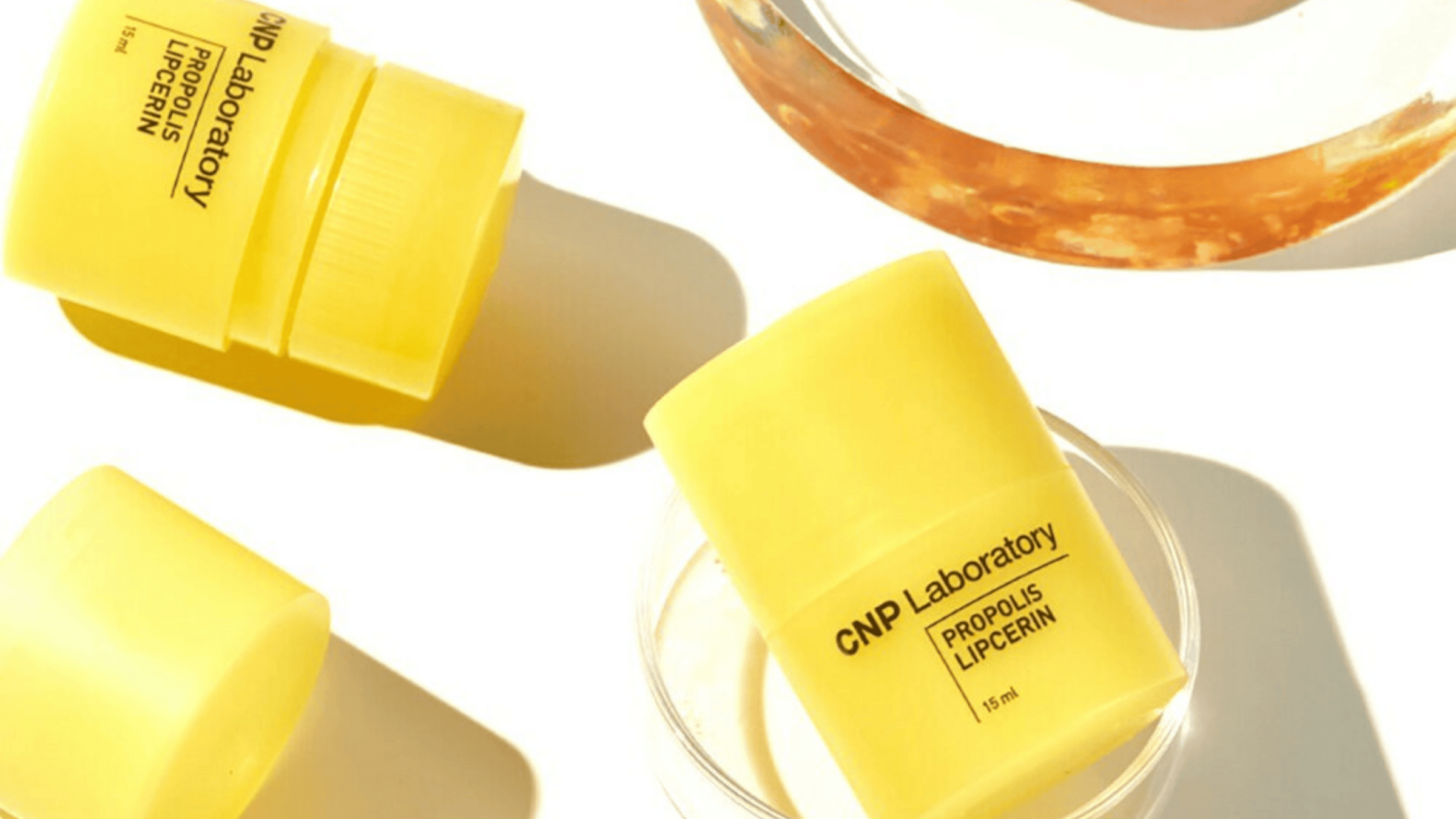 Bilden visar CNP Laboratory Propolis Lipcerin i gula tuber bredvid en glaskruka med honung. Produkten, avsedd för läppvård, framhävs av den livliga gula färgen och mjuka bakgrunden.