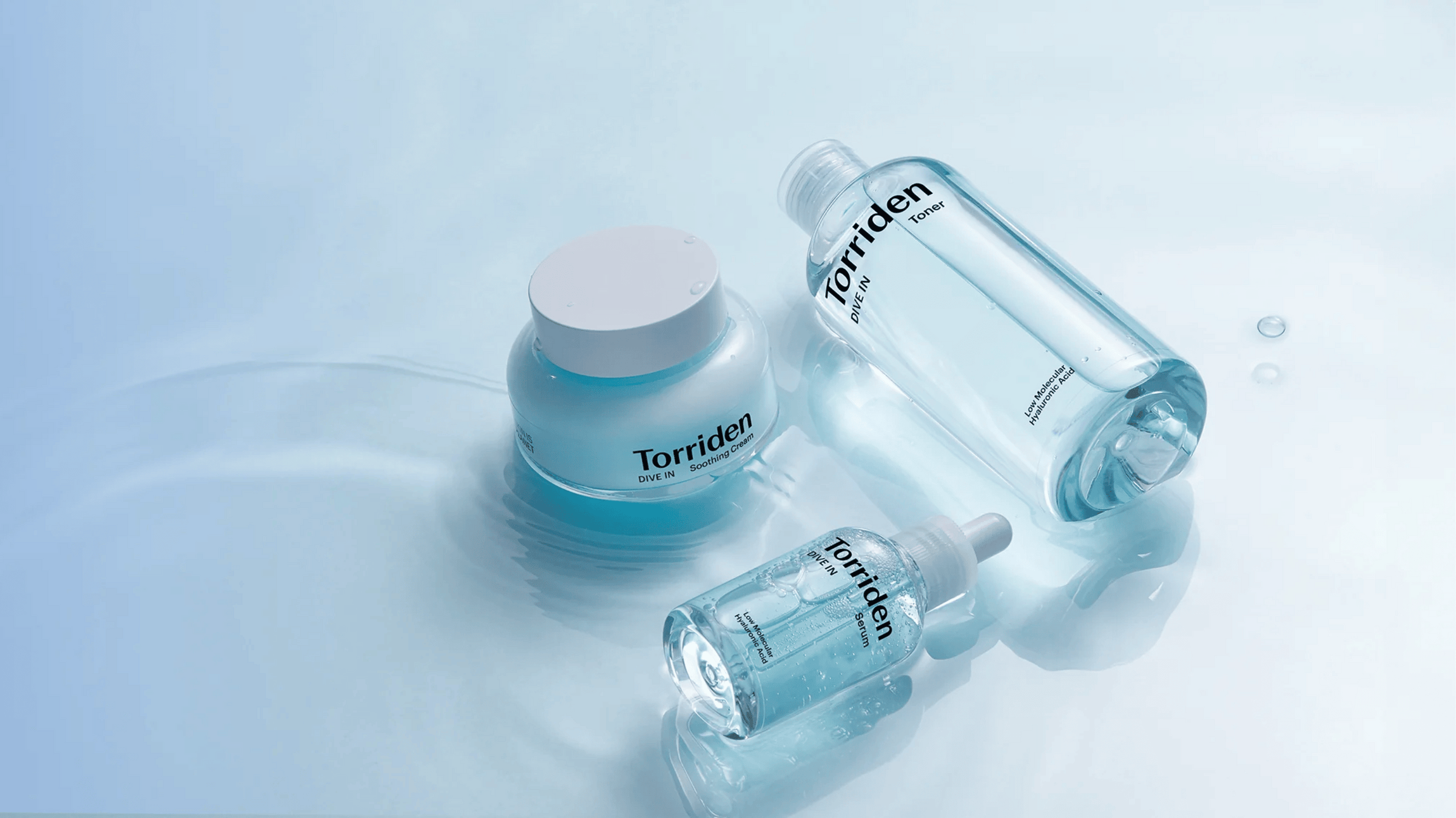 En trio av Torriden DIVE IN-produkter: toner, serum och soothing cream, visas på en vattenliknande yta. De ljusblå förpackningarna förstärker känslan av återfuktning och fräschör, med fokus på hyaluronsyrans intensiva fuktgivande egenskaper.