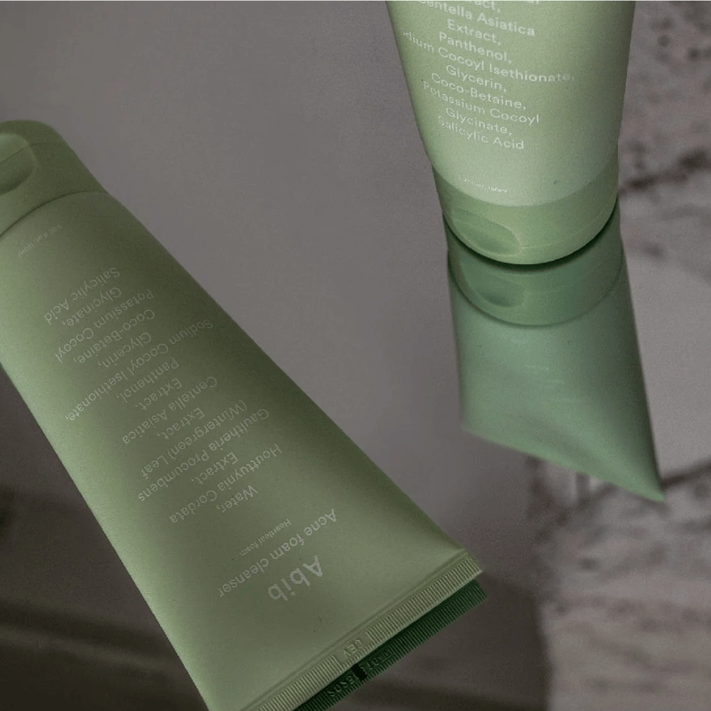 Grön tub av Abib Acne Foam Cleanser Heartleaf liggande på ett spegelbord med produktens framsida och text speglad. Ingredienser som salicylsyra, centella och hjärtbladsextrakt syns tydligt på förpackningen.