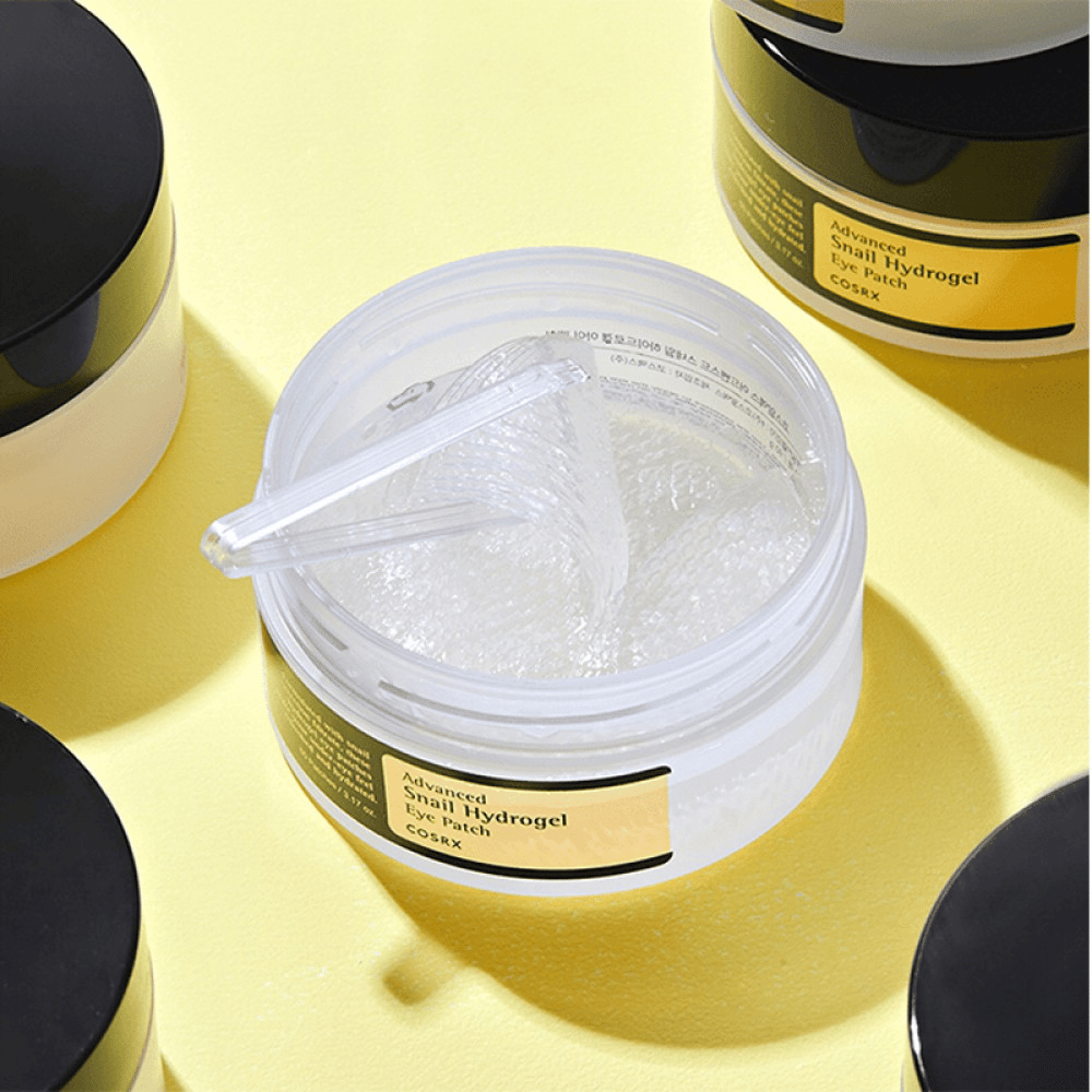 En öppen burk COSRX Advanced Snail Hydrogel Eye Patch med genomskinliga ögonpatches och en liten spatel, omgiven av fler stängda burkar på gul bakgrund.