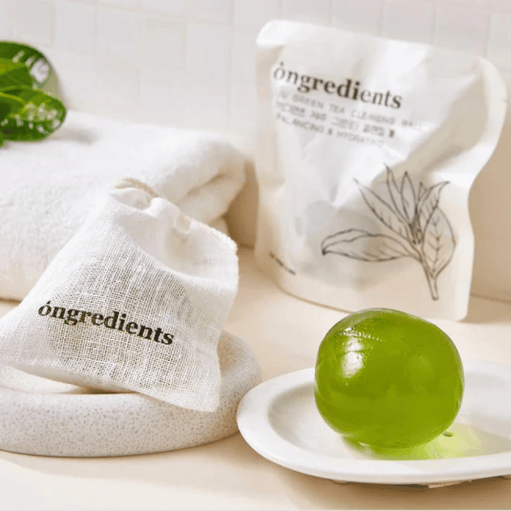 Óngredients Jeju Green Tea Cleansing Ball placerad på fat med förpackning och handduk – skonsam rengöring med grönt te från Jeju.