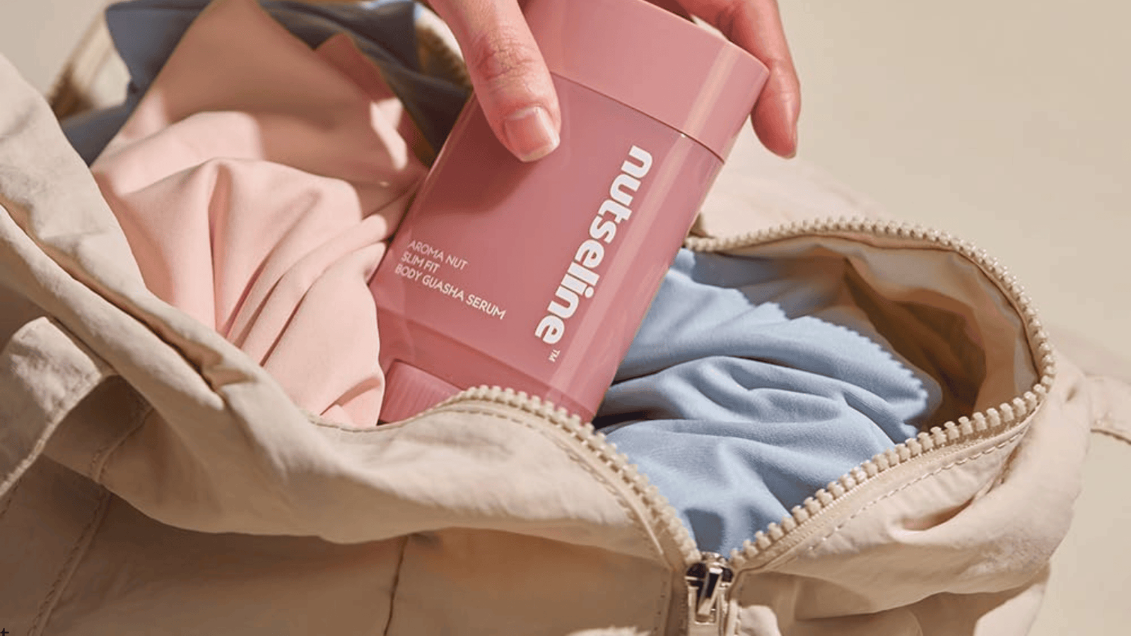 En hand placerar en rosa kroppsserumprodukt från Nuseline i en öppen tränings- eller weekendväska fylld med kläder. Bilden förmedlar en praktisk och stilren känsla med fokus på enkel hudvård och kroppsvård att ta med på resan eller till gymmet.