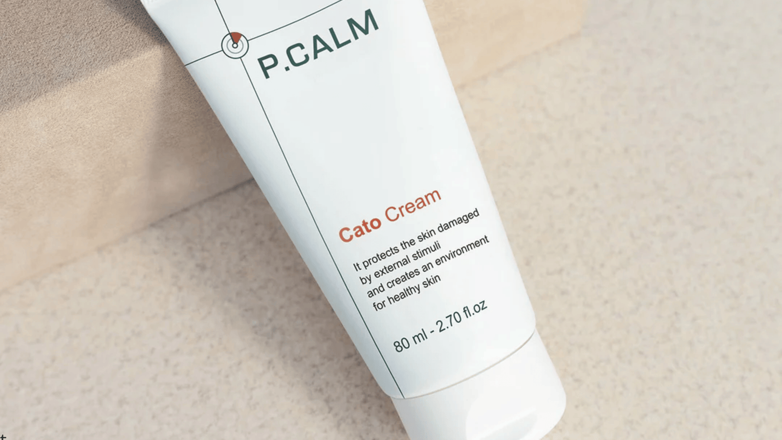 Närbild på en vit tub från P.CALM med produkten Cato Cream placerad på en ljus, neutral yta. Designen är minimalistisk med gröna och orange detaljer, och fokus ligger på hudvårdskrämen samt dess lugnande och skyddande egenskaper för känslig hud.