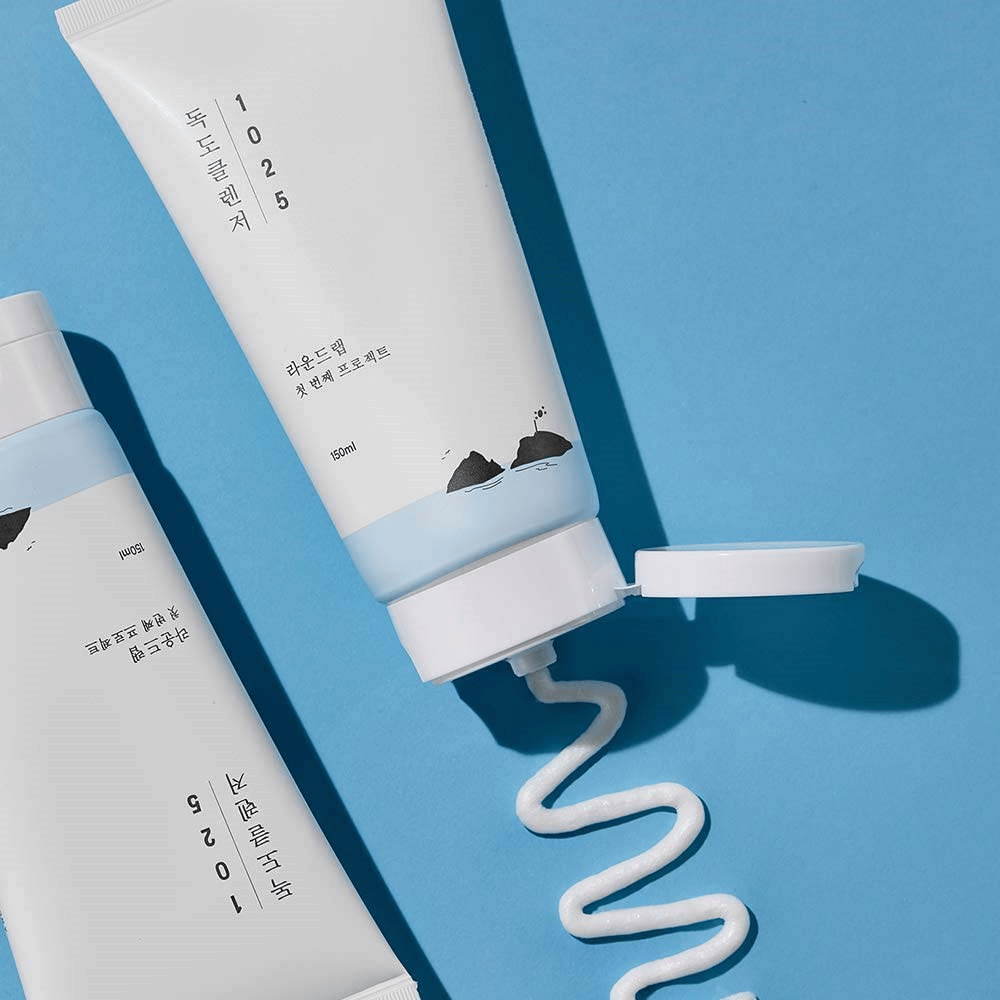 Δύο σωλήνες από 1025 Dokdo Cleanser σε μπλε φόντο, εκ των οποίων ο ένας με ανοιχτό καπάκι και μια κυματιστή λωρίδα καθαρισμού πιεσμένη.