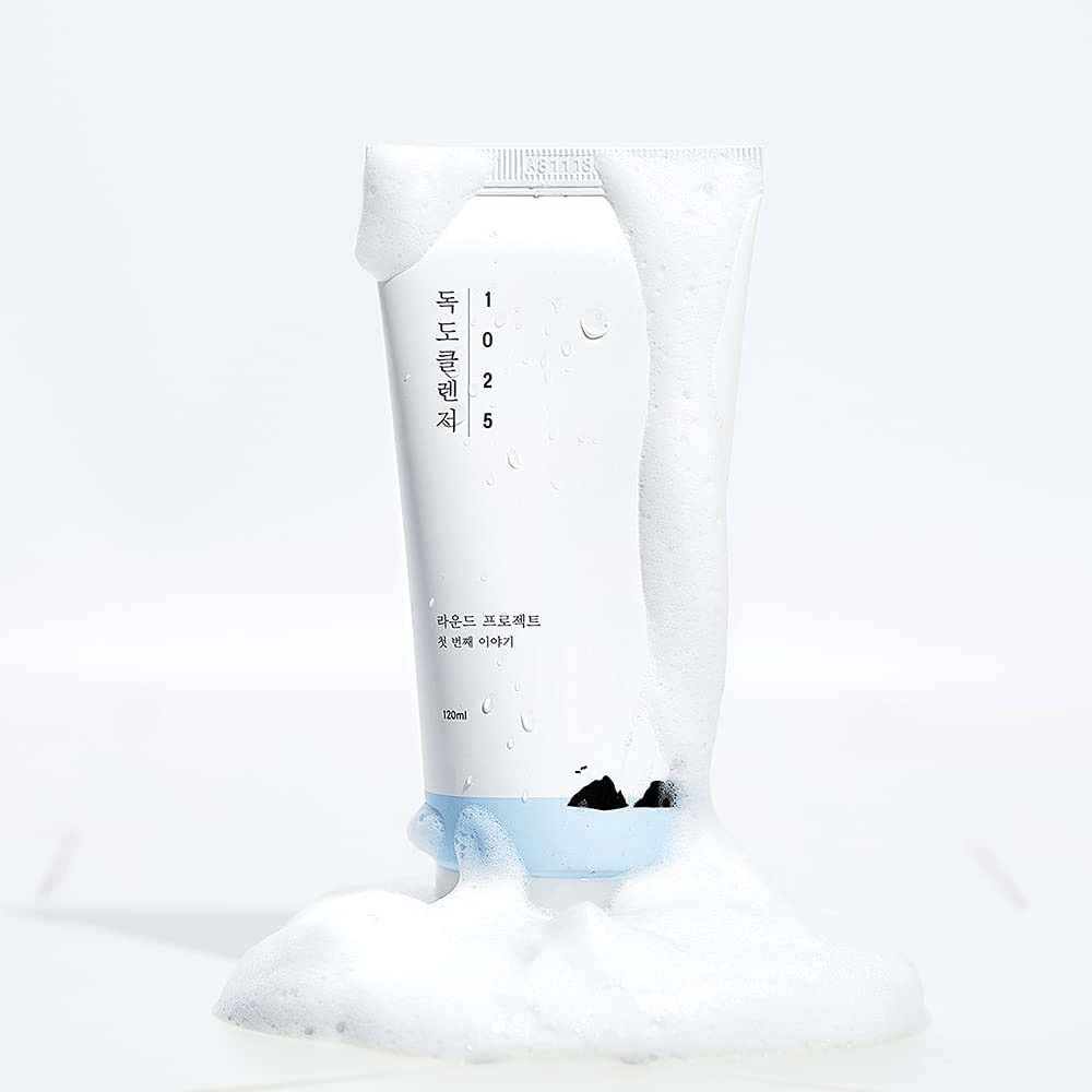 1025 Dokdo Cleanser σε λευκό σωλήνα καλυμμένο με αφρό, με σταγόνες νερού και αφρό γύρω από τη βάση, απομονωμένο σε ανοιχτό φόντο.