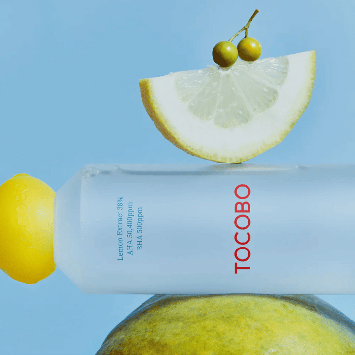 Flaska med TOCOBO AHA BHA Lemon Toner, dekorerad med ett lock i form av en citron. En citronskiva och små gröna bär balanserar ovanpå flaskan mot en blå bakgrund.
