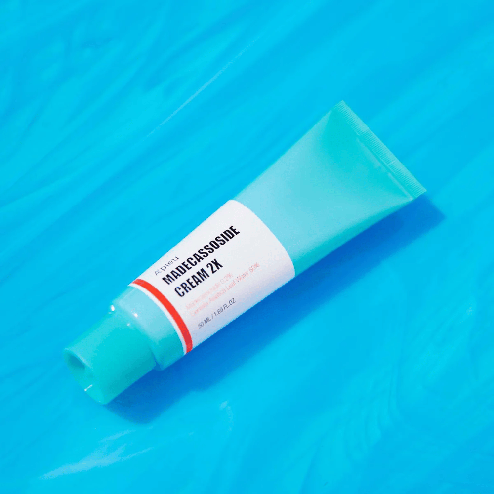Ένα σωληνάριο A'PIEU Madecassoside Cream 2X βρίσκεται διαγώνια πάνω σε ένα μπλε φόντο με ρυτίδες που μοιάζουν με νερό. Ο σωλήνας έχει λευκό σώμα με ανοιχτό τιρκουάζ καπάκι και ετικέτα χρωματιστή από κόκκινο σε πορτοκαλί όπου το όνομα του προϊόντος και το ενεργό συστατικό Madecassoside 2% είναι ξεκάθαρα γραμμένο. Στην ετικέτα αναγράφεται επίσης η ποσότητα που είναι 50 ml / 1.8 fl oz. Η φωτεινή και πολύχρωμη παρουσίαση δίνει μια φρέσκια και ενεργητική αίσθηση.