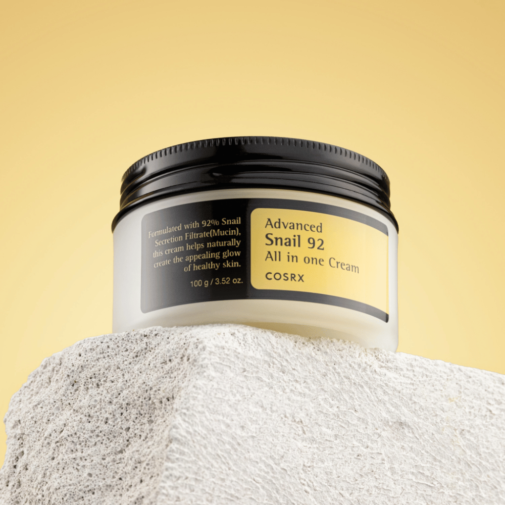 Ένα βάζο του COSRX Advanced Snail 92 All in one Cream τοποθετημένο σε βραχώδη επιφάνεια με κίτρινο φόντο.