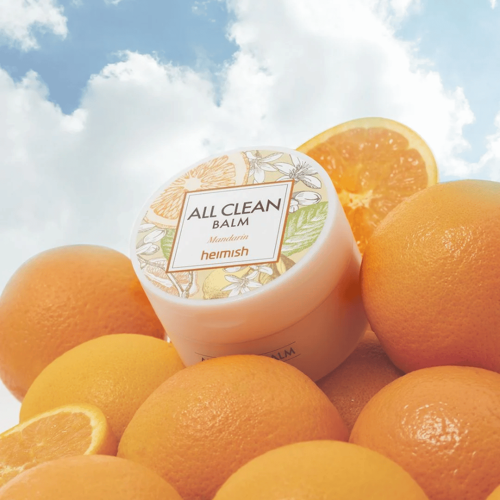Βάζο από Heimish All Clean Balm Mandarin τοποθετημένο σε ένα σωρό με πορτοκάλια, με έναν μπλε ουρανό στο φόντο.