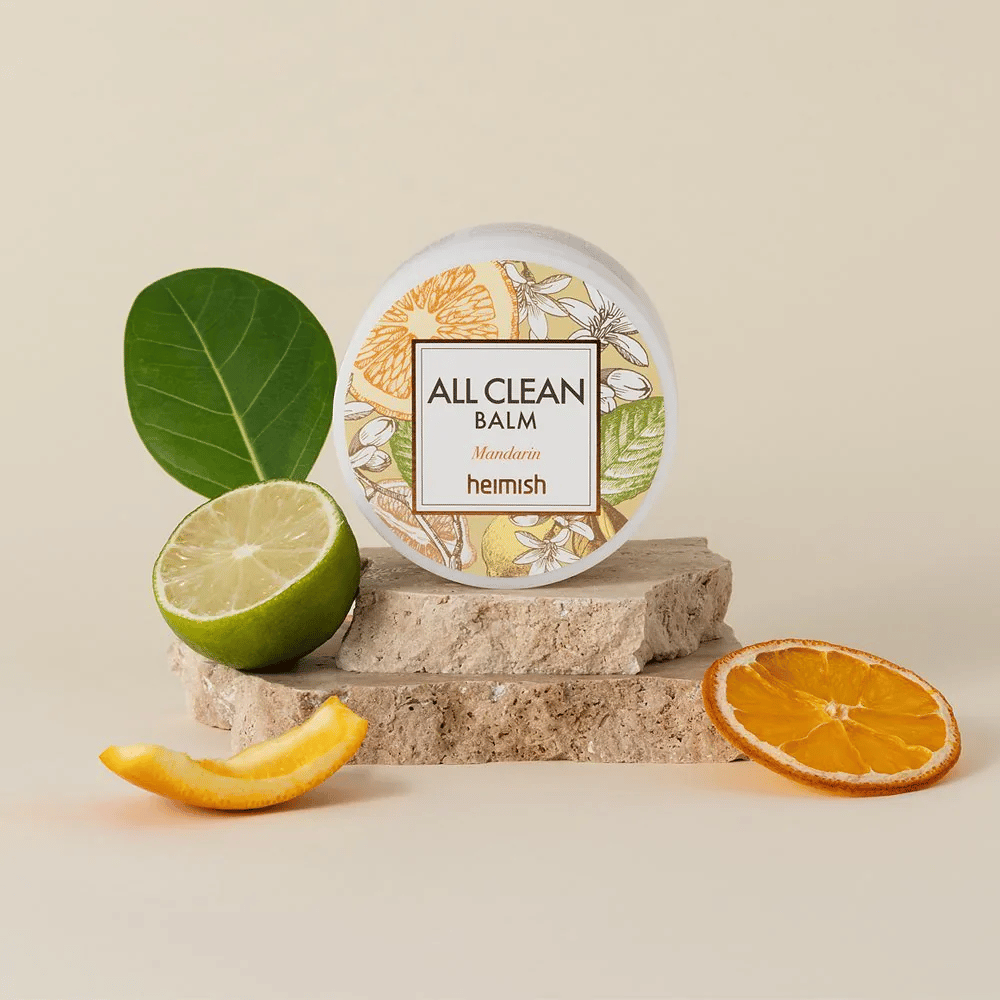 Βάζο από Heimish All Clean Balm Mandarin τοποθετημένο σε πέτρες, περιτριγυρισμένο από φλούδες μανταρινιού και φύλλα εσπεριδοειδών, μπροστά από ένα φωτεινό φόντο.