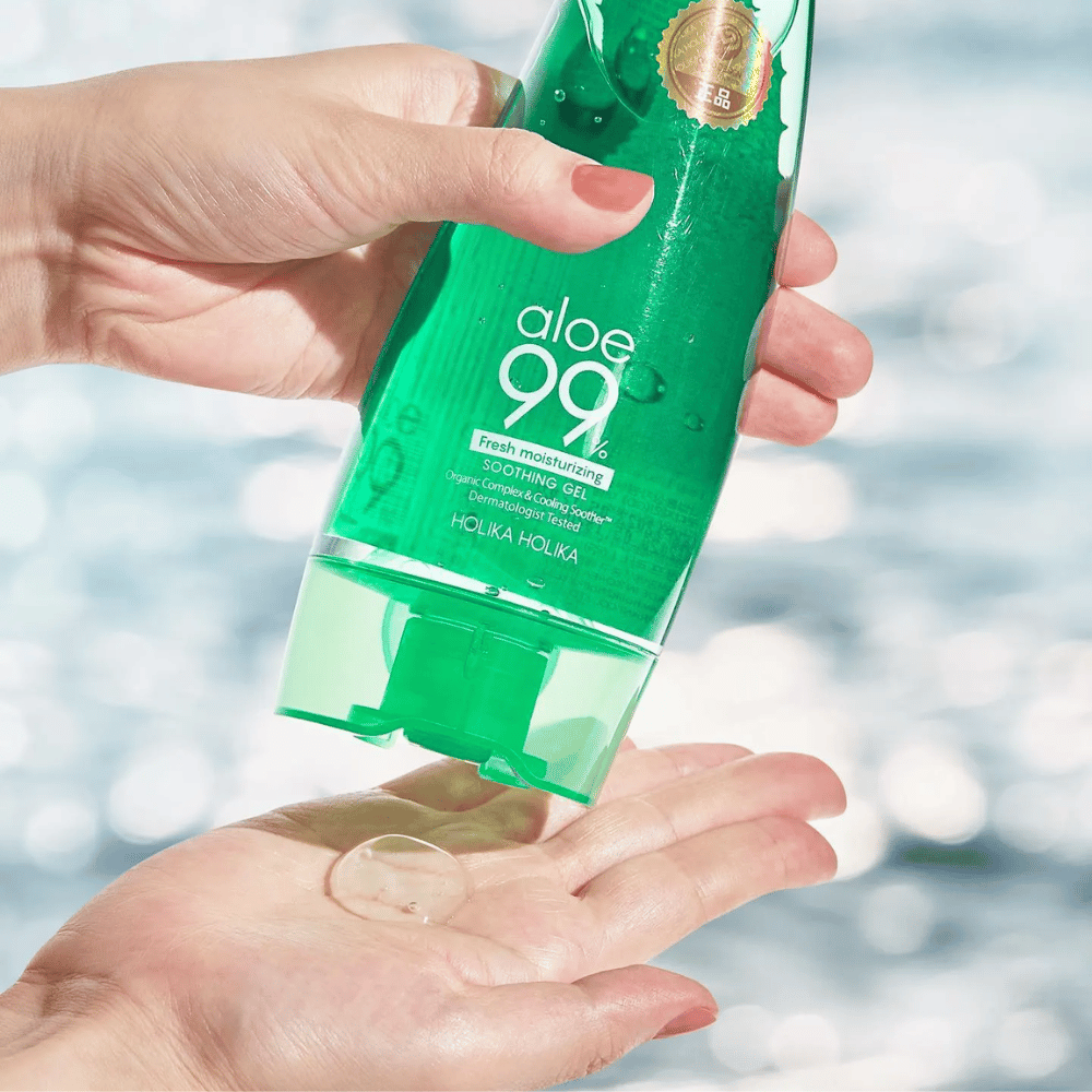 Ένα χέρι κρατάει ένα πράσινο σωληνάριο με Holika Holika Aloe 99% Soothing Gel, ενώ ένα άλλο χέρι πιάνει μια σταγόνα τζελ που εκτοξεύεται. Το φόντο είναι θολό και δείχνει ένα υδατώδες, λαμπερό μοτίβο.
