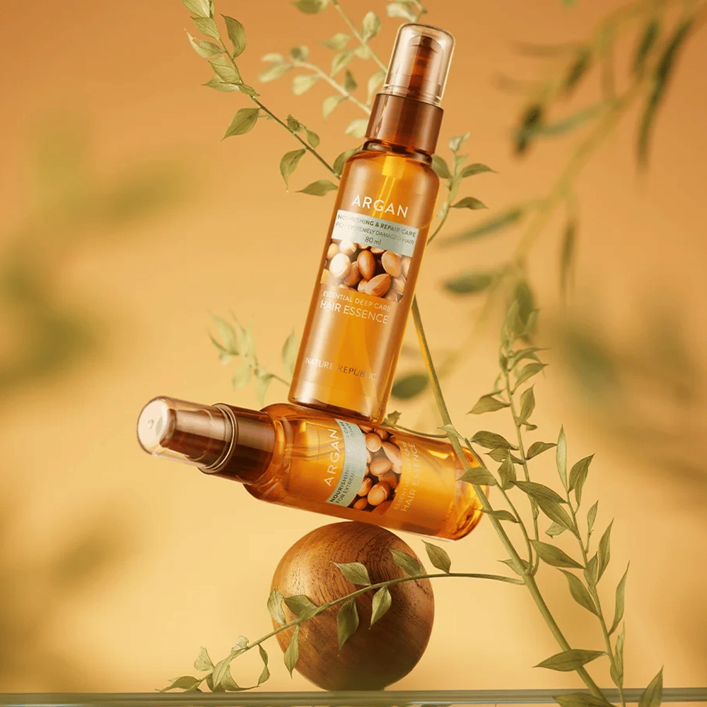 Δύο μπουκάλια Nature Republic Argan Essential Moisture Hair Oil τοποθετημένα ανάμεσα σε πράσινα φύλλα – μεταξένια λάμψη για ξηρά μαλλιά.