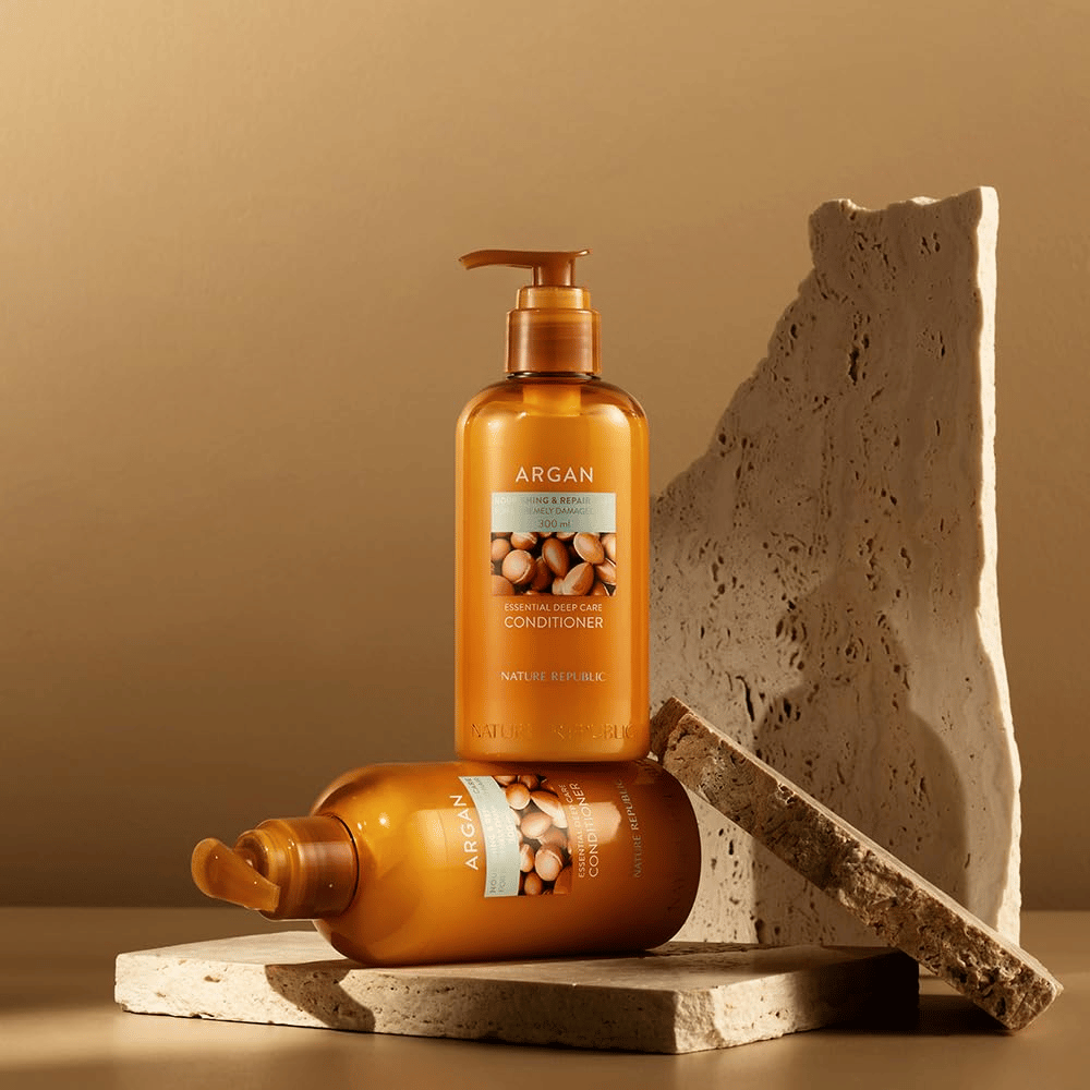 Δύο μπουκάλια από Nature Republic Argan Essential Moisture Repairing Conditioner σε πέτρινη επιφάνεια σε ζεστό τόνο.