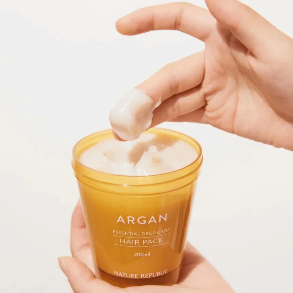 Χέρι παίρνει τη κρεμώδη φόρμουλα από Nature Republic Argan Essential Moisture Repairing Hair Mask – εντατική θρέψη για ξηρά μαλλιά.