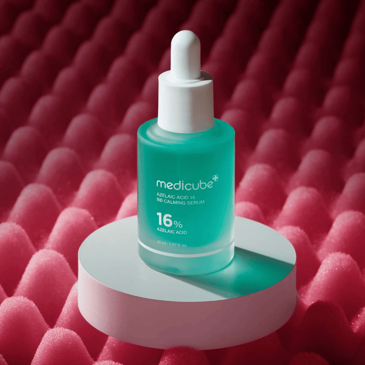Εικόνα προϊόντος του Medicube Azelaic Acid 16 BB Calming Serum τοποθετημένου σε λευκή επιφάνεια βάθρου με ροζ υφή στο φόντο που αναδεικνύει το καταπραϋντικό και βελτιωτικό για την επιδερμίδα προφίλ του ορού.