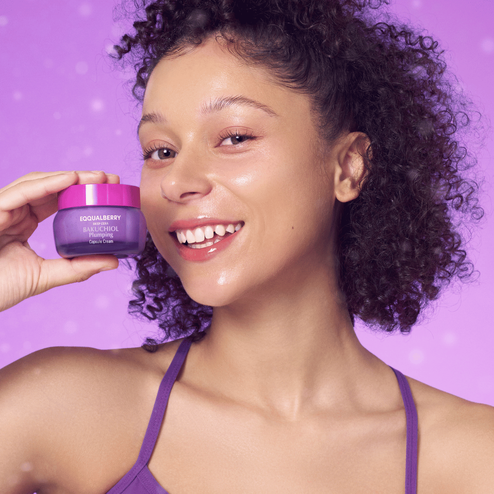 Γυναίκα με λαμπερή επιδερμίδα κρατάει EQQUALBERRY Bakuchiol Plumping Capsule Cream και χαμογελάει μπροστά σε μωβ φόντο.