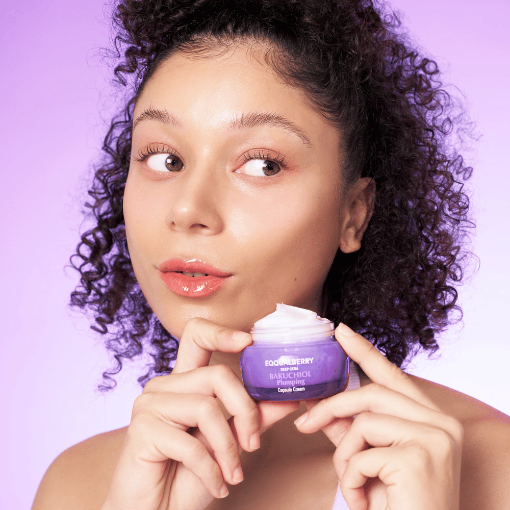 Γυναίκα κρατάει EQQUALBERRY Bakuchiol Plumping Capsule Cream στο χέρι της και δείχνει το ενυδατωμένο, λαμπερό δέρμα της μπροστά σε μωβ φόντο.