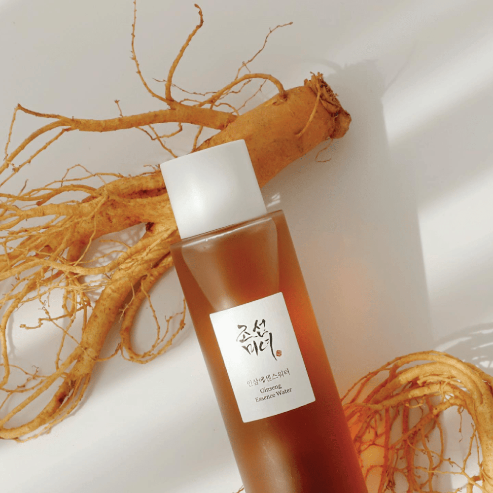 Ένα καφέ μπουκάλι Beauty of Joseon Ginseng Essence Water με λευκό πώμα βρίσκεται δίπλα σε μια φρέσκια ρίζα ginseng σε λευκό φόντο.