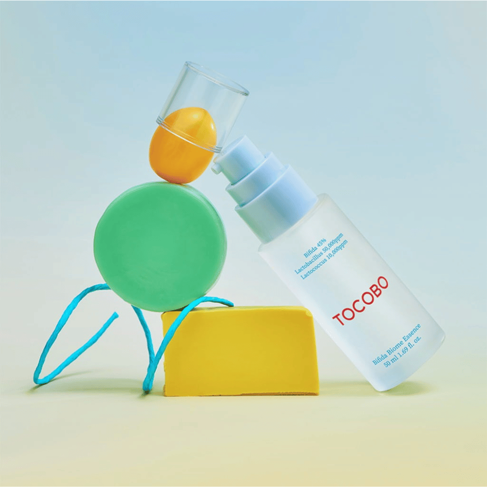 TOCOBO Bifida Biome Essence σε παγωμένο μπουκάλι αντλίας με κόκκινο λογότυπο, τοποθετημένο σε πολύχρωμο φόντο με πράσινα, κίτρινα και πορτοκαλί γεωμετρικά σχήματα. Το κείμενο στη φιάλη αναφέρει Bifida 5%, Lactobacillus 3 000 ppm και Lactococcus 1 000 ppm.