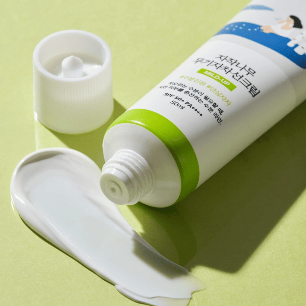 Κοντινό πλάνο του Birch Juice Moisturizing Mild Up Sunscreen με ξεβιδωμένο καπάκι, λευκή αντηλιακή κρέμα απλωμένη σε ανοιχτό πράσινο φόντο, SPF 50+ PA++++, 50 ml.