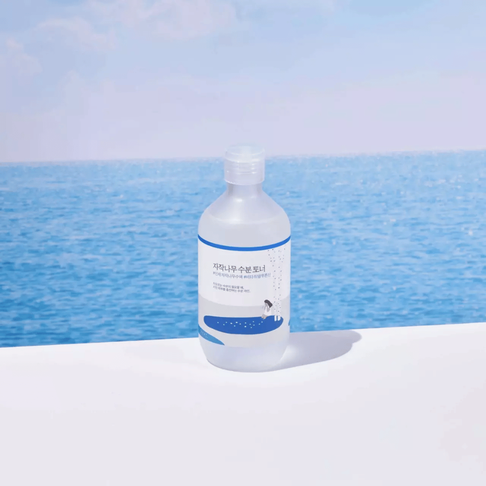 Μπουκάλι ROUND LAB Birch Juice Moisturizing Toner στέκεται σε λευκή επιφάνεια με θάλασσα και γαλάζιο ουρανό στο φόντο