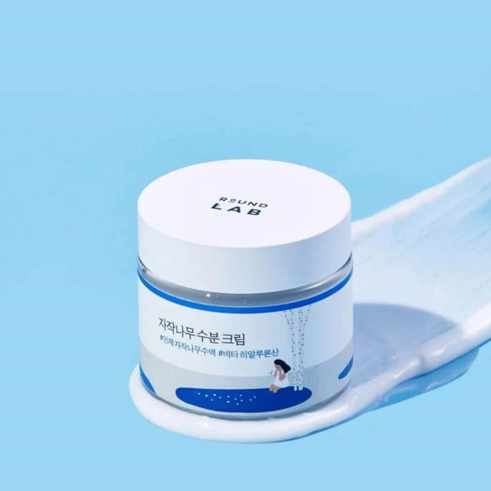 Round Lab Birch Juice Moisturizing Cream σε μπλε απόχρωση βάζο με λευκό καπάκι, τοποθετημένο σε στρώμα κρέμας με μπλε φόντο.