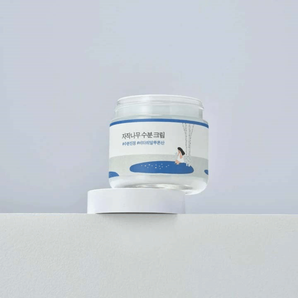 Round Lab Birch Juice Moisturizing Cream σε διαφανές βάζο με λευκή ετικέτα και μπλε εικονογράφηση γυναίκας δίπλα σε σημύδα, τοποθετημένο σε λευκή επιφάνεια με ανοιχτό φόντο.