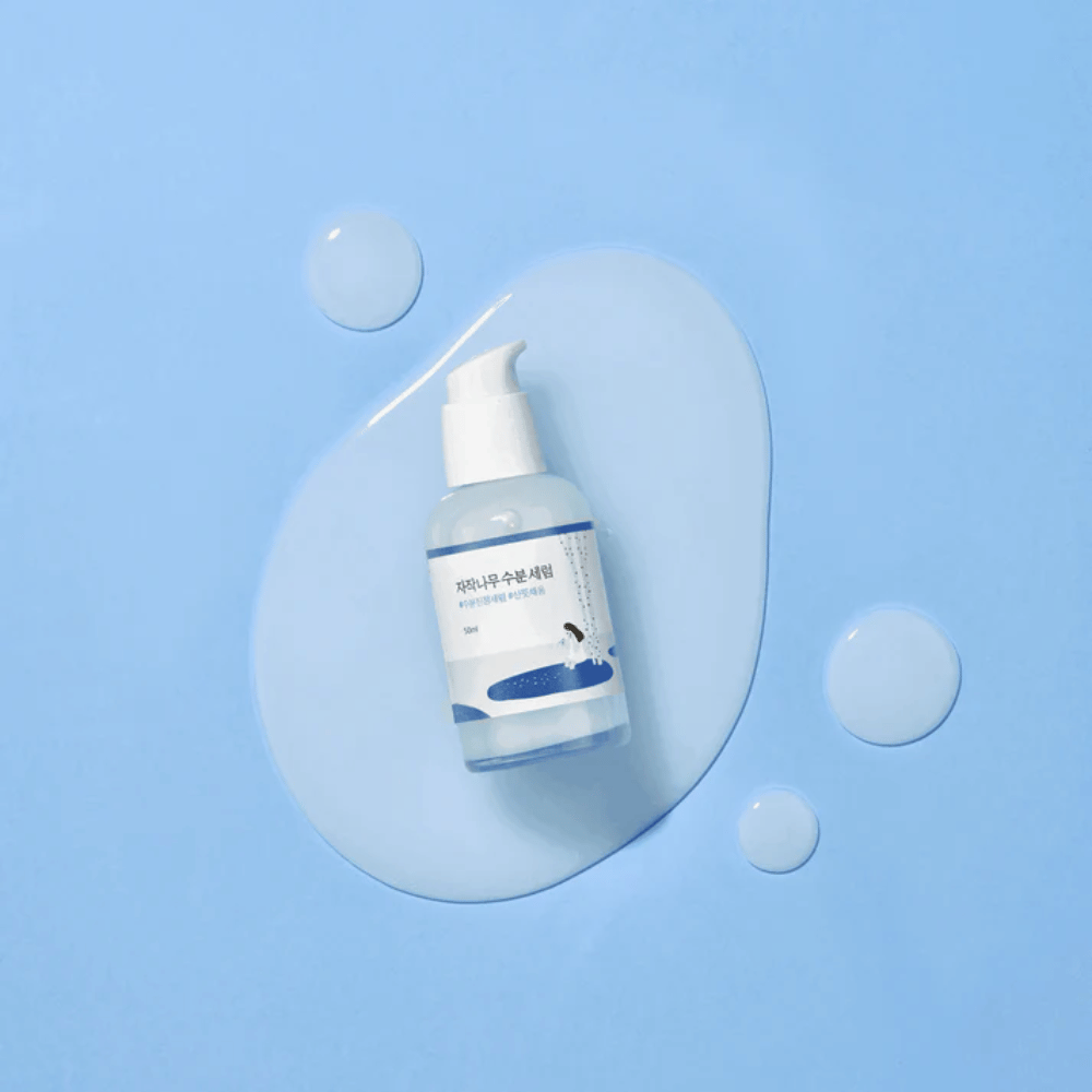 Round Lab Birch Juice Moisturizing Serum σε διαφανές μπουκάλι αντλίας τοποθετημένο σε μπλε φόντο με σταγόνες και χυμένο ορό.