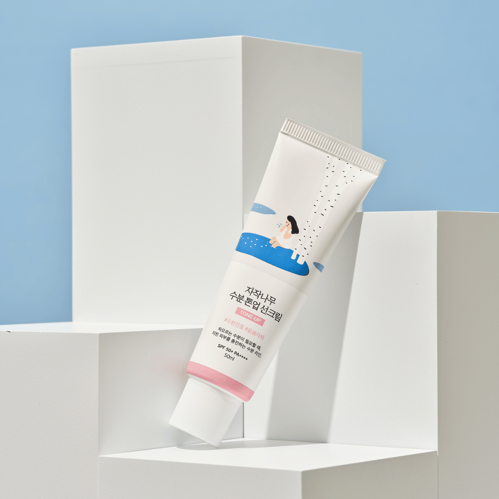 Ένα σωληνάριο με Birch Juice Moisturizing Tone Up Sunscreen τοποθετημένο σε λευκούς κύβους με φόντο ανοιχτό μπλε. Το σωληνάριο είναι λευκό με μια εικονογράφηση μιας γυναίκας δίπλα σε μια σημύδα και νερό. Το κείμενο στα κορεατικά αναφέρει ότι το προϊόν έχει SPF 50+ PA++++ και περιέχει 50 ml.