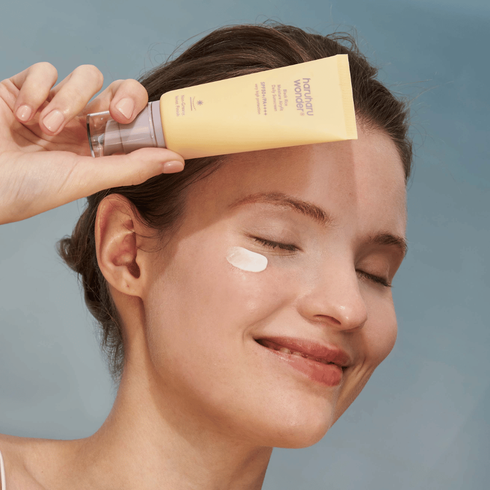 Μια χαμογελαστή γυναίκα εφαρμόζει Black Rice Moisture Airyfit Daily Sunscreen SPF 50+ PA++++ από Haruharu Wonder στο πρόσωπό της. Κρατάει ένα κίτρινο σωληνάριο με αντηλιακή κρέμα και έχει μια σταγόνα από το προϊόν στο μάγουλό της. Το φόντο είναι ανοιχτό μπλε.