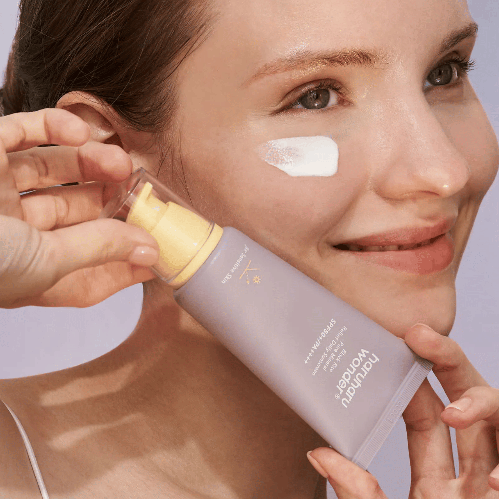 Μια χαμογελαστή γυναίκα εφαρμόζει Haruharu Wonder Black Rice Pure Mineral Relief Daily Sunscreen SPF 50+ PA++++ στο πρόσωπό της. Κρατάει έναν μοβ σωλήνα με αντηλιακή κρέμα και έχει μια σταγόνα από το προϊόν στο μάγουλό της. Το φόντο είναι ανοιχτό μοβ.