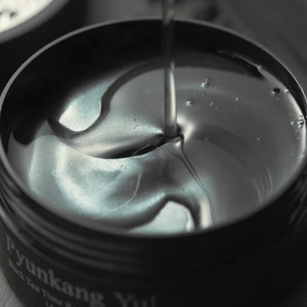 Κοντινό πλάνο σε μαύρα hydrogel eye patches μέσα σε ανοιχτό βαζάκι από την Pyunkang Yul. Τα γυαλιστερά patches βρίσκονται μέσα σε serum και αναδεικνύουν την ενυδατική και πολυτελή αίσθηση του προϊόντος.