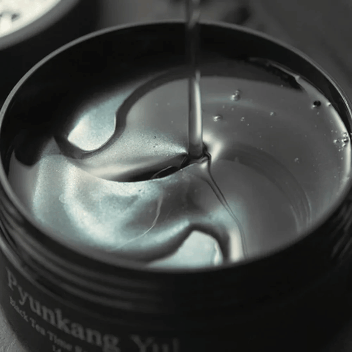Κοντινό πλάνο σε μαύρα hydrogel eye patches μέσα σε ανοιχτό βαζάκι από την Pyunkang Yul. Τα γυαλιστερά patches βρίσκονται μέσα σε serum και αναδεικνύουν την ενυδατική και πολυτελή αίσθηση του προϊόντος.