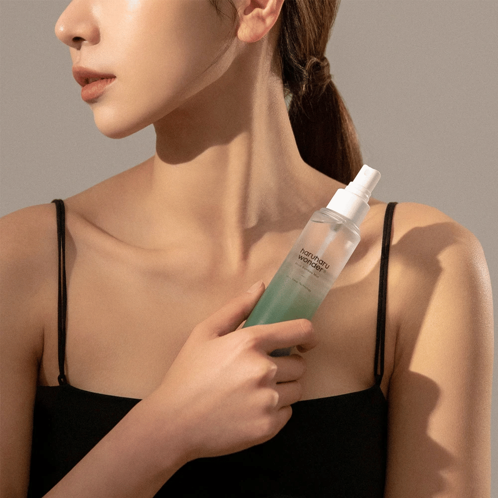 Εικόνα του HaruHaru Wonder Black Bamboo Mist που κρατιέται από μια γυναίκα. Το προϊόν είναι σε ένα διάφανο μπουκάλι σπρέι με πράσινη απόχρωση και λευκή αντλία. Η γυναίκα έχει μια χαλαρή εμφάνιση, φοράει μια μαύρη μπλούζα και εστιάζει στη χρήση του σπρέι για να δώσει στο δέρμα υγρασία και λάμψη. Το μπουκάλι είναι μέρος μιας ρουτίνας περιποίησης δέρματος.