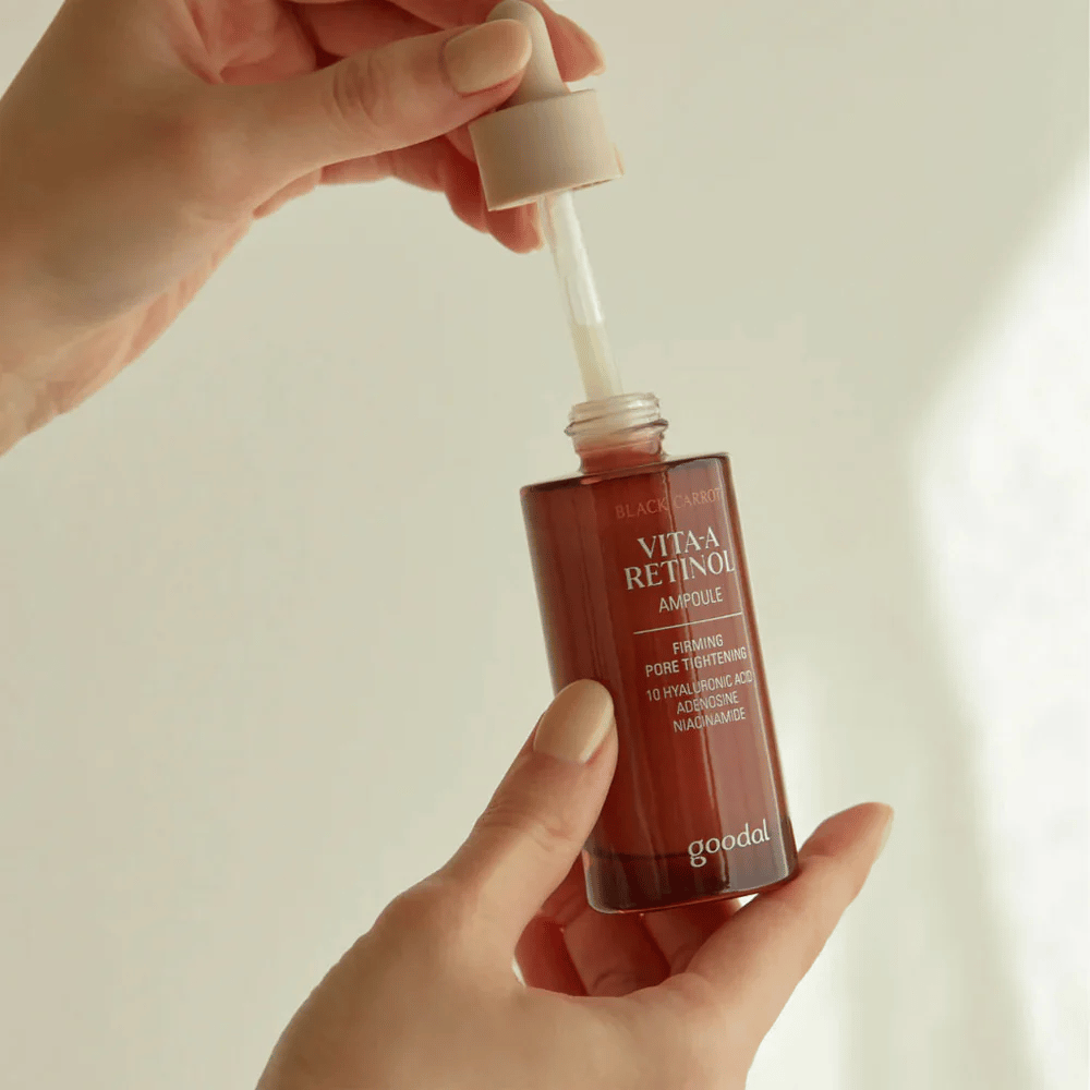 Ένα χέρι κρατάει ένα μπουκάλι με Goodal Black Carrot Vita-A Retinol Firming Ampoule. Η ετικέτα του μπουκαλιού δείχνει το όνομα και τις ιδιότητες του προϊόντος, συμπεριλαμβανομένων των "Σύσφιξη Πόρων" και συστατικών όπως 10% υαλουρονικό οξύ, αδενοσίνη και νιασιναμίδη. Η πιπέτα είναι σε μπεζ χρώμα.