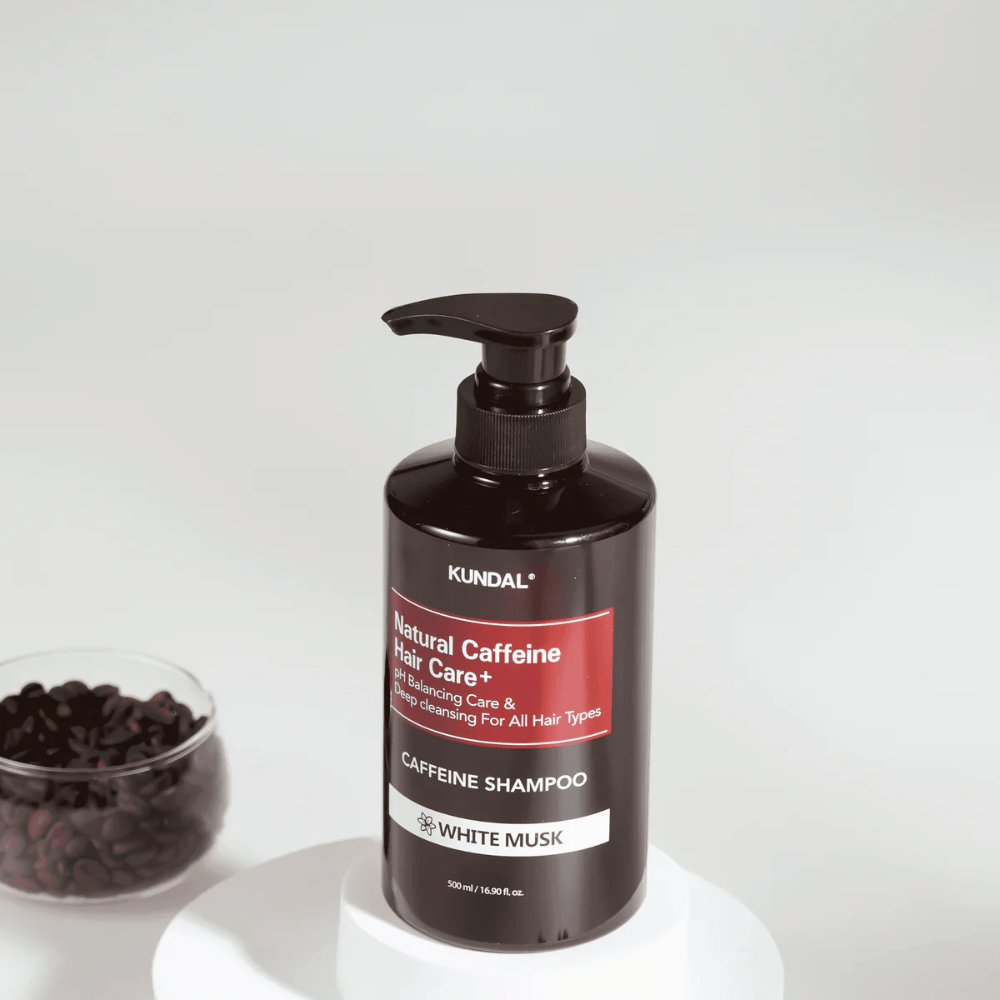 KUNDAL Natural Caffeine Hair Care+ Shampoo White Musk σε μαύρο μπουκάλι με αντλία, 500 ml, τοποθετημένο δίπλα σε ένα μπολ με κόκκους καφέ.