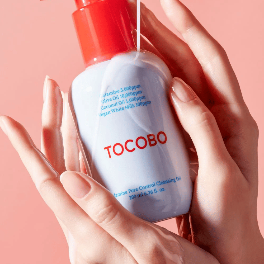 TOCOBO Calamine Pore Control Cleansing Oil κρατιέται στα χέρια πάνω σε ροζ φόντο, η κόκκινη αντλία του μπουκαλιού και η λίστα συστατικών φαίνονται καθαρά.