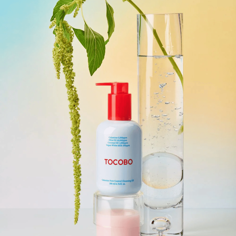 TOCOBO Calamine Pore Control Cleansing Oil με κόκκινη αντλία στέκεται δίπλα σε ένα ψηλό γυάλινο σωλήνα γεμάτο με νερό και ένα πράσινο φυτό, πάνω σε ανοιχτό παστέλ φόντο.