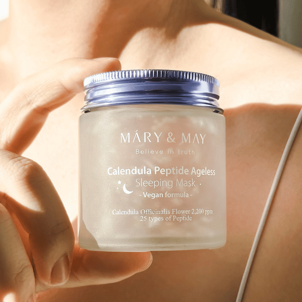 Μια γυναίκα κρατάει ένα βαζάκι με MARY & MAY Calendula Peptide Ageless Sleeping Mask. Το βαζάκι είναι διάφανο με μοβ καπάκι και το κείμενο "Calendula Peptide Ageless Sleeping Mask" φαίνεται καθαρά. Το προϊόν είναι μια βίγκαν μάσκα ύπνου με καλέντουλα και πεπτίδια, σχεδιασμένη για να δώσει στο δέρμα μια νεανική λάμψη κατά τη διάρκεια της νύχτας.