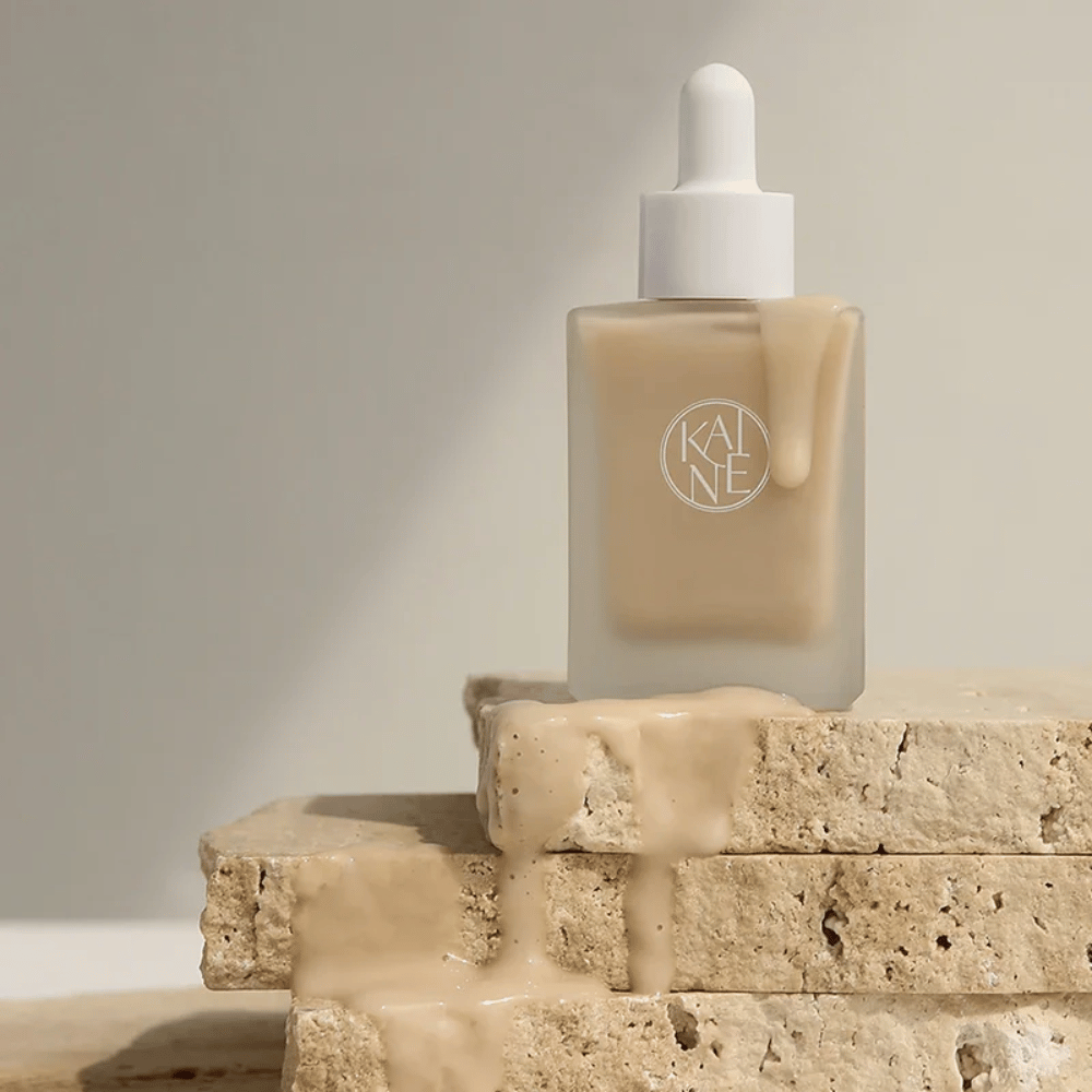 KANE Chaga Collagen Charging Serum σε ένα παγωμένο γυάλινο μπουκάλι με πιπέτα. Κρεμώδης υφή ρέει πάνω από φυσικές πέτρες σε απαλό και μινιμαλιστικό φως.