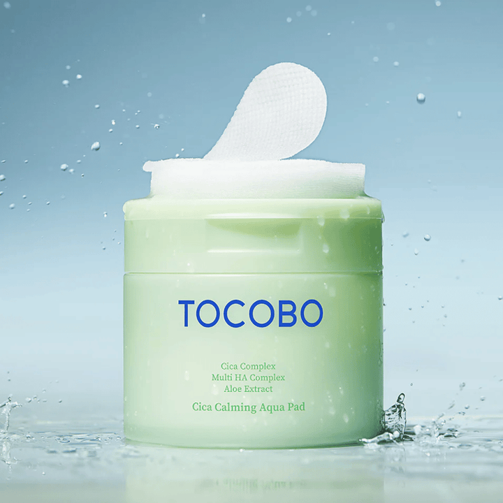 Ανοιχτό βαζάκι με TOCOBO Cica Calming Aqua Pad όπου ένα toner pad προεξέχει από τη συσκευασία. Σταγόνες νερού γύρω από το προϊόν ενισχύουν την αίσθηση έντονης ενυδάτωσης και φρεσκάδας.
