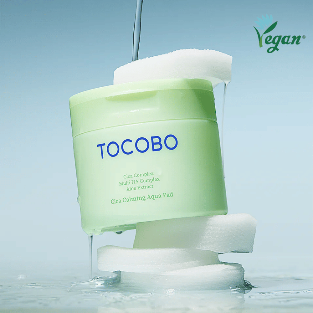Το TOCOBO Cica Calming Aqua Pad τοποθετημένο ανάμεσα σε toner pads με essence που ρέει. Το σύμβολο vegan τονίζει τη vegan σύνθεση του προϊόντος και το φιλικό προς το δέρμα προφίλ του.