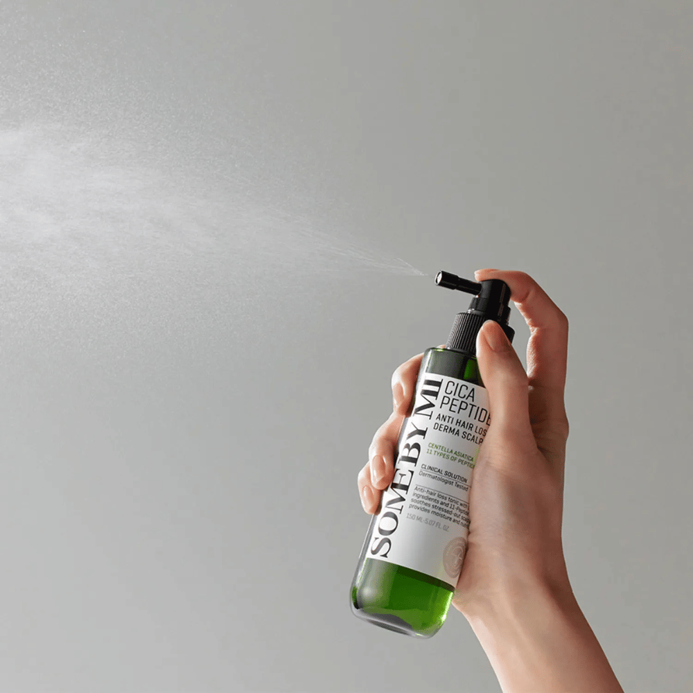 Χέρι που ψεκάζει SOME BY MI Cica Peptide Anti Hair Loss Derma Scalp Tonic από ένα πράσινο μπουκάλι με μαύρο ακροφύσιο και λευκή ετικέτα σε γκρι φόντο.