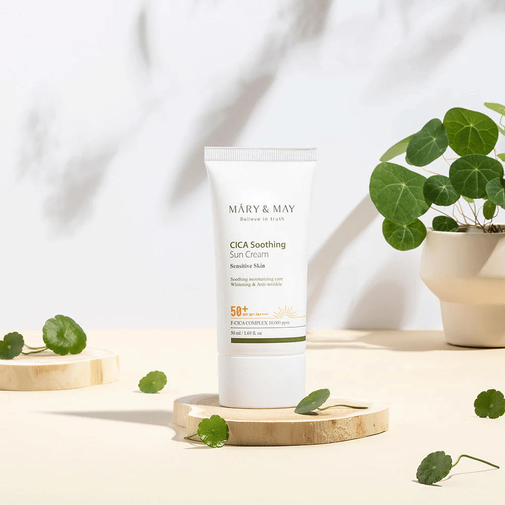 Το Mary&May CICA Soothing Sun Cream τοποθετημένο σε ξύλινο βάθρο με πράσινα φύλλα centella γύρω του. Η εικόνα τονίζει τα καταπραϋντικά συστατικά για το δέρμα και τη φυσική έμπνευση πίσω από το προϊόν.