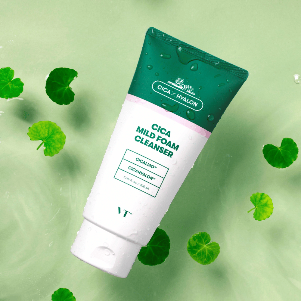 VT Cica Mild Foam Cleanser ξαπλωμένο σε ανοιχτό πράσινο υπόστρωμα με σταγόνες νερού στη συσκευασία και περιτριγυρισμένο από πράσινα φύλλα centella asiatica, που υπογραμμίζουν τις καταπραϋντικές ιδιότητες του προϊόντος.