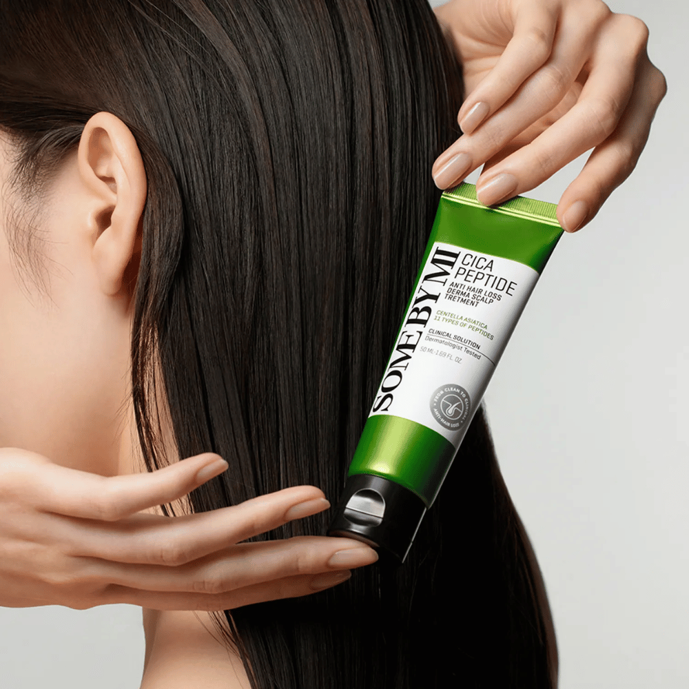 Γυναίκα εφαρμόζει SOME BY MI Cica Peptide Anti Hair Loss Derma Scalp Treatment σε μακριά, σκούρα μαλλιά με πράσινο και λευκό σωλήνα.