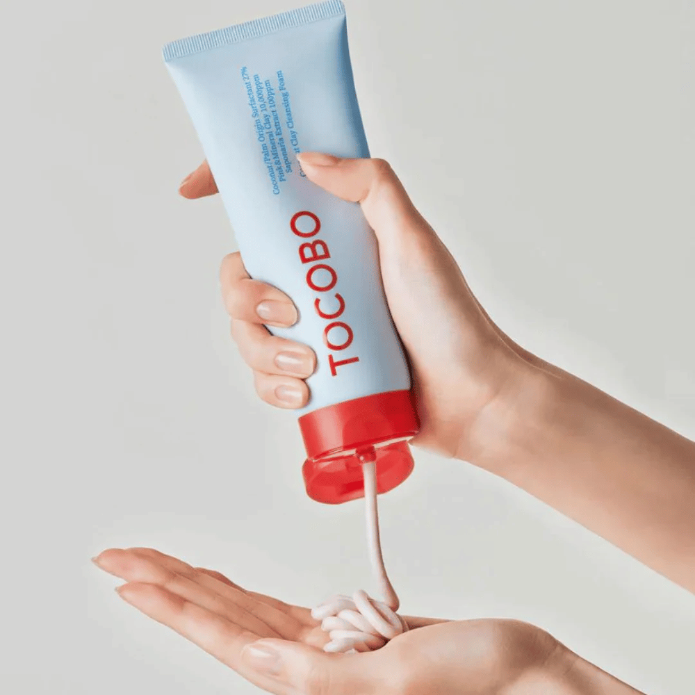 Ένα άτομο κρατά έναν σωλήνα του TOCOBO Coconut Clay Cleansing Foam. Ο σωλήνας είναι ανοιχτό μπλε με κόκκινο καπάκι και το κείμενο "TOCOBO" στο μπροστινό μέρος. Το προϊόν πιέζεται έξω από τον σωλήνα και σχηματίζεται σαν σαμπουάν στο χέρι. Το φόντο είναι ουδέτερο και δίνει έμφαση στο προϊόν.