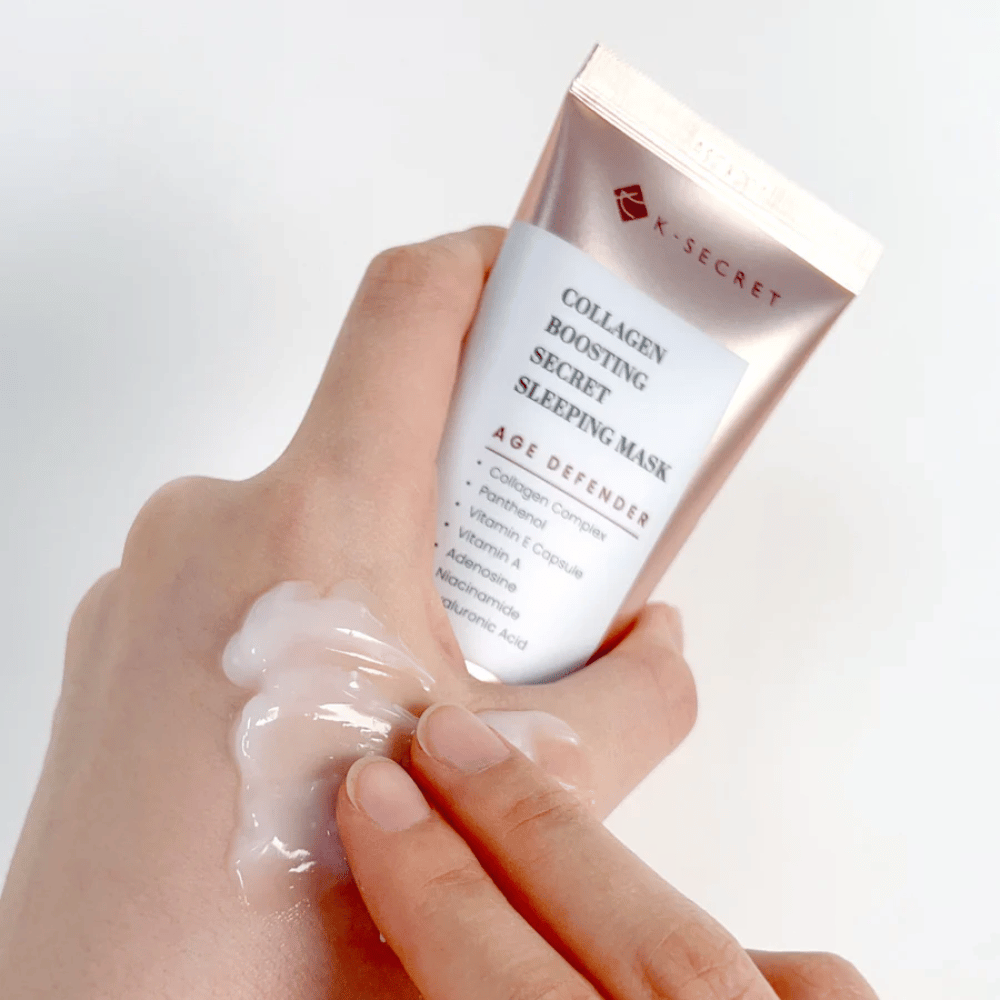 Ένα χέρι κρατάει το Collagen Boosting Secret Sleeping Mask ενώ το άλλο εφαρμόζει μια γενναιόδωρη ποσότητα κρέμας στην άνω πλευρά του χεριού - δείχνει την υφή και την εφαρμογή του προϊόντος.