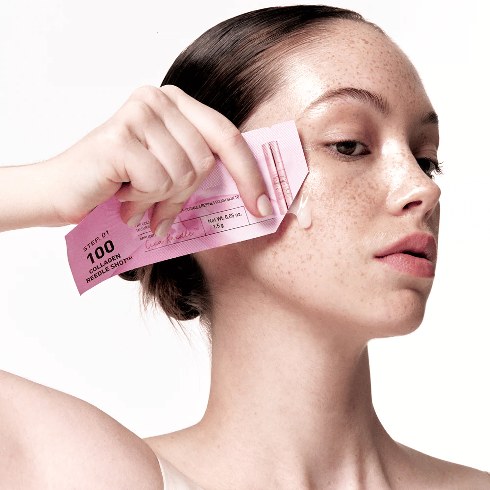 Γυναίκα εφαρμόζει το βήμα 1 του Collagen Reedle Shot 100 με μια ροζ συσκευασία μιας χρήσης στο μάγουλο. Μια καθαρή γέλη φαίνεται στο δέρμα, υποδεικνύοντας το πρώτο βήμα της διπλής φάσης περιποίησης δέρματος.