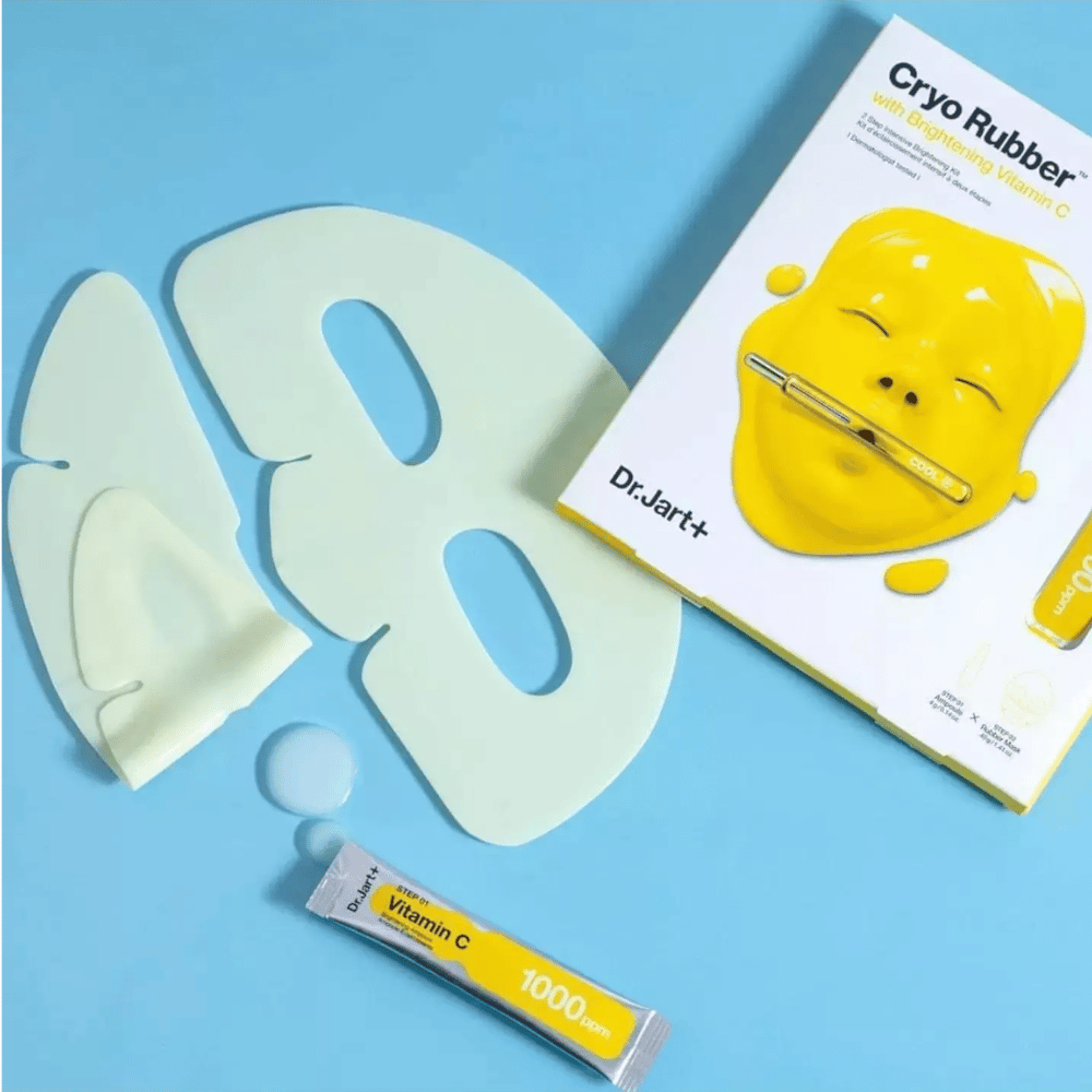 Εικόνα του Dr.Jart+'s 'Cryo Rubber™ με Φωτεινή Βιταμίνη C' προϊόν ομορφιάς. Σε ένα μπλε φόντο φαίνεται μια λευκή μάσκα προσώπου από καουτσούκ, ένα μικρό σωληνάριο με την ένδειξη 'Βιταμίνη C 1000ppm', και ένα κουτί με το όνομα του προϊόντος και εικόνα της κίτρινης μάσκας.